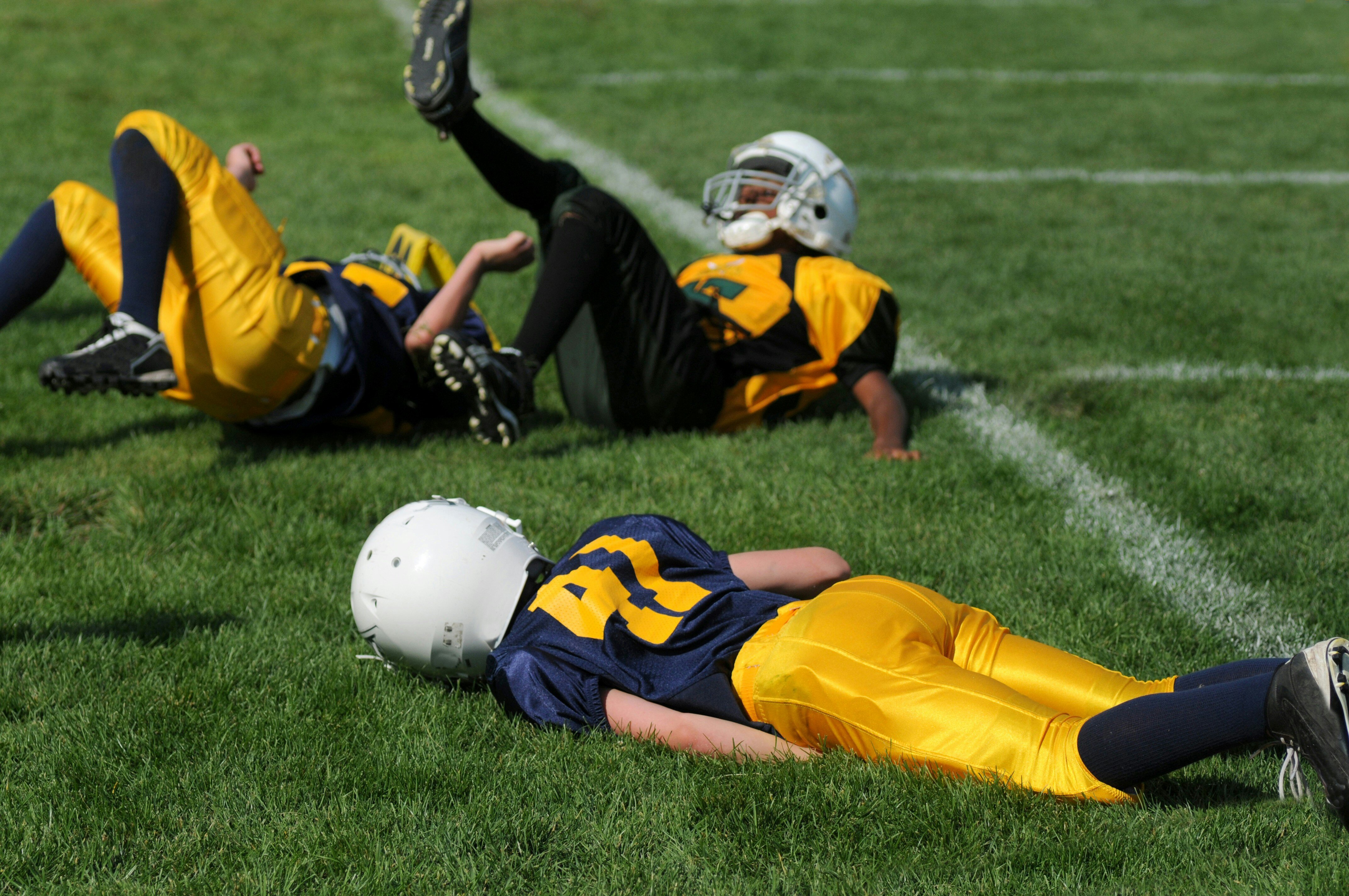 The image shows three young football players wearing helmets and uniforms lying on the ground during what appears to be a youth football game or practice. The players are dressed in bright yellow and dark blue uniforms. Two players are lying on their backs, while the third has their legs in the air, as if falling or recovering from a tackle. The grass field is visible, and they seem to be taking a moment to rest or recover after a play.