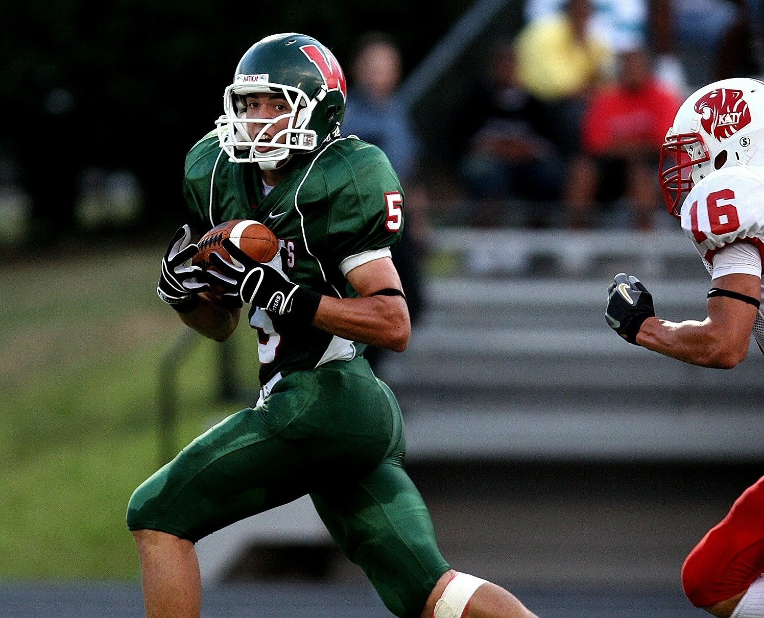 A football player wearing a green uniform is running with the ball, looking ahead as he is pursued by another player in a red and white uniform.